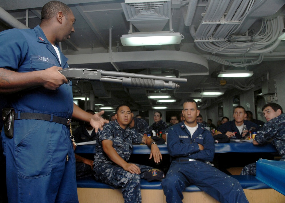 A U.S. Navy petty officer named Fairley is conducting a weapons familiarization or safety briefing aboard a naval vessel, holding a pump-action shotgun while addressing a group of seated sailors. The audience is wearing standard U.S. Navy blue digital camouflage (NWU Type I) uniforms and appears attentive. The setting is an interior compartment of a ship, likely a berthing or mess area.