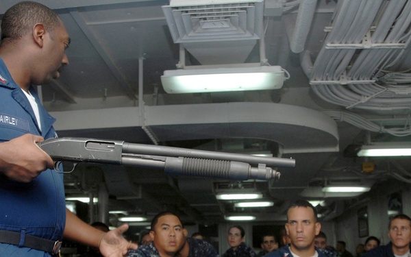 A U.S. Navy petty officer named Fairley is conducting a weapons familiarization or safety briefing aboard a naval vessel, holding a pump-action shotgun while addressing a group of seated sailors. The audience is wearing standard U.S. Navy blue digital camouflage (NWU Type I) uniforms and appears attentive. The setting is an interior compartment of a ship, likely a berthing or mess area.