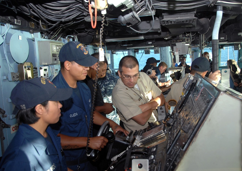 The image shows military personnel on the bridge of a naval vessel, with sailors in both dark blue and khaki uniforms gathered around navigation and control consoles. A person in khaki uniform, likely an officer, appears to be demonstrating or explaining equipment operation to others. The setting is clearly an active ship's bridge with visible ocean through the windows.