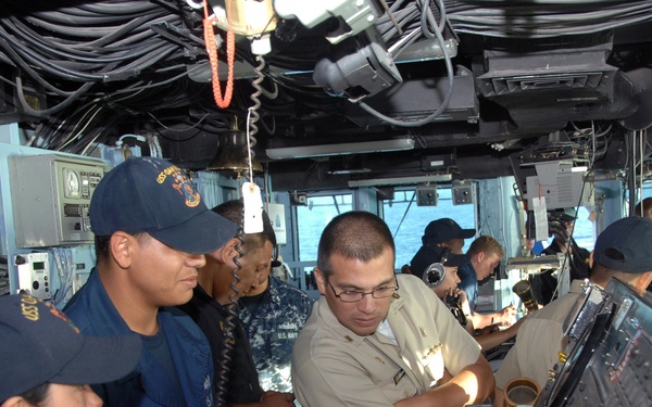 The image shows military personnel on the bridge of a naval vessel, with sailors in both dark blue and khaki uniforms gathered around navigation and control consoles. A person in khaki uniform, likely an officer, appears to be demonstrating or explaining equipment operation to others. The setting is clearly an active ship's bridge with visible ocean through the windows.