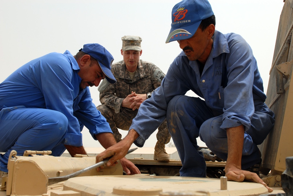 Three men are working on what appears to be a military vehicle in a desert environment. Two men wearing blue work uniforms and baseball caps are performing hands-on mechanical work on the vehicle's surface, while a U.S. Army soldier in camouflage uniform observes and supervises. The scene suggests a training or advisory interaction between military personnel and civilian workers.