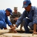 Three men are working on what appears to be a military vehicle in a desert environment. Two men wearing blue work uniforms and baseball caps are performing hands-on mechanical work on the vehicle's surface, while a U.S. Army soldier in camouflage uniform observes and supervises. The scene suggests a training or advisory interaction between military personnel and civilian workers.