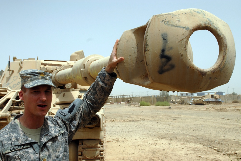 A U.S. Army soldier in ACU (Army Combat Uniform) is holding and examining the muzzle brake of a large artillery piece or tank cannon in what appears to be a military base in a desert environment. The soldier's name tape reads 'DELON' and he appears to be explaining or demonstrating the weapon component. The setting shows a sandy, arid landscape consistent with Middle Eastern deployment.