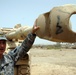 A U.S. Army soldier in ACU (Army Combat Uniform) is holding and examining the muzzle brake of a large artillery piece or tank cannon in what appears to be a military base in a desert environment. The soldier's name tape reads 'DELON' and he appears to be explaining or demonstrating the weapon component. The setting shows a sandy, arid landscape consistent with Middle Eastern deployment.