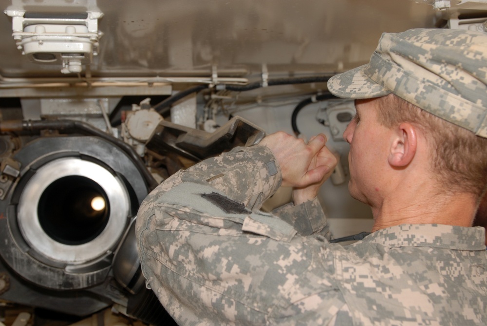 A U.S. Army soldier in Army Combat Uniform (ACU) digital camouflage pattern is performing maintenance or inspection work on a military vehicle, likely a tank or armored fighting vehicle. The soldier is working in a confined space beneath or alongside the vehicle's hull, manipulating a component with both hands. A large circular opening, possibly a gun barrel bore or exhaust port, is visible in the foreground.
