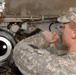 A U.S. Army soldier in Army Combat Uniform (ACU) digital camouflage pattern is performing maintenance or inspection work on a military vehicle, likely a tank or armored fighting vehicle. The soldier is working in a confined space beneath or alongside the vehicle's hull, manipulating a component with both hands. A large circular opening, possibly a gun barrel bore or exhaust port, is visible in the foreground.