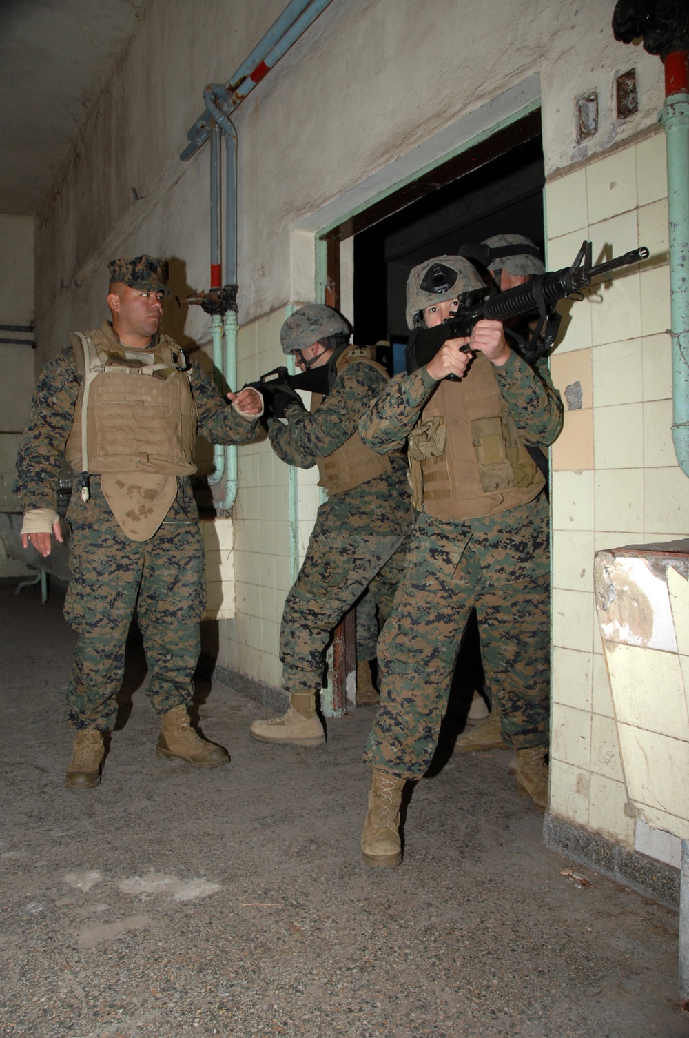 The image shows a group of U.S. Marines in MARPAT digital camouflage uniforms conducting military urban operations training, specifically a room-clearing or building-entry drill. Personnel are equipped with body armor, helmets, and M16/M4-style rifles in a tactically ready position near a doorway. The setting appears to be a deteriorated building used as a training facility.
