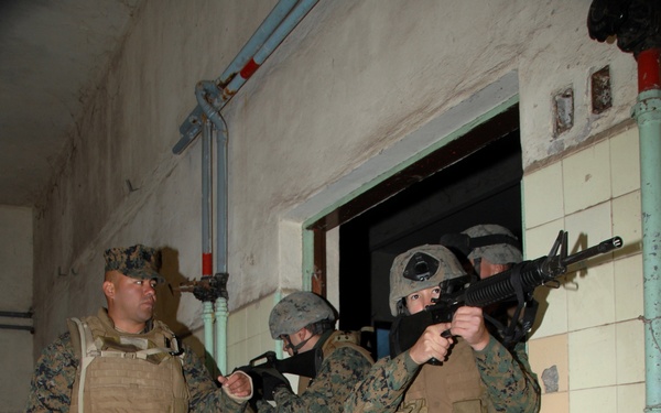 The image shows a group of U.S. Marines in MARPAT digital camouflage uniforms conducting military urban operations training, specifically a room-clearing or building-entry drill. Personnel are equipped with body armor, helmets, and M16/M4-style rifles in a tactically ready position near a doorway. The setting appears to be a deteriorated building used as a training facility.