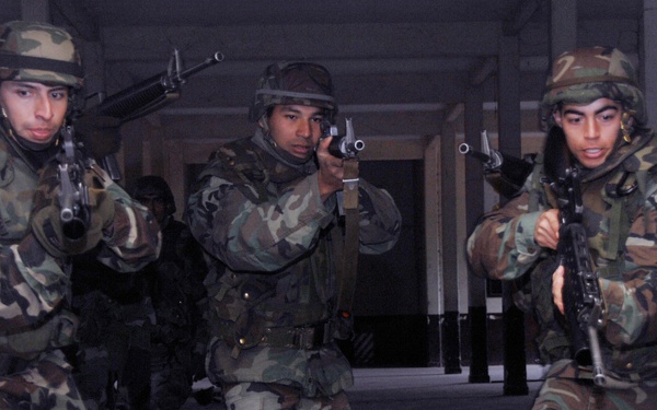 Three soldiers in woodland camouflage uniforms and helmets are conducting a building clearing or urban warfare training exercise, moving through a doorway in tactical formation. Each soldier is armed and has weapons raised in a ready position. Additional personnel are visible in the background of what appears to be an indoor training facility or abandoned building.
