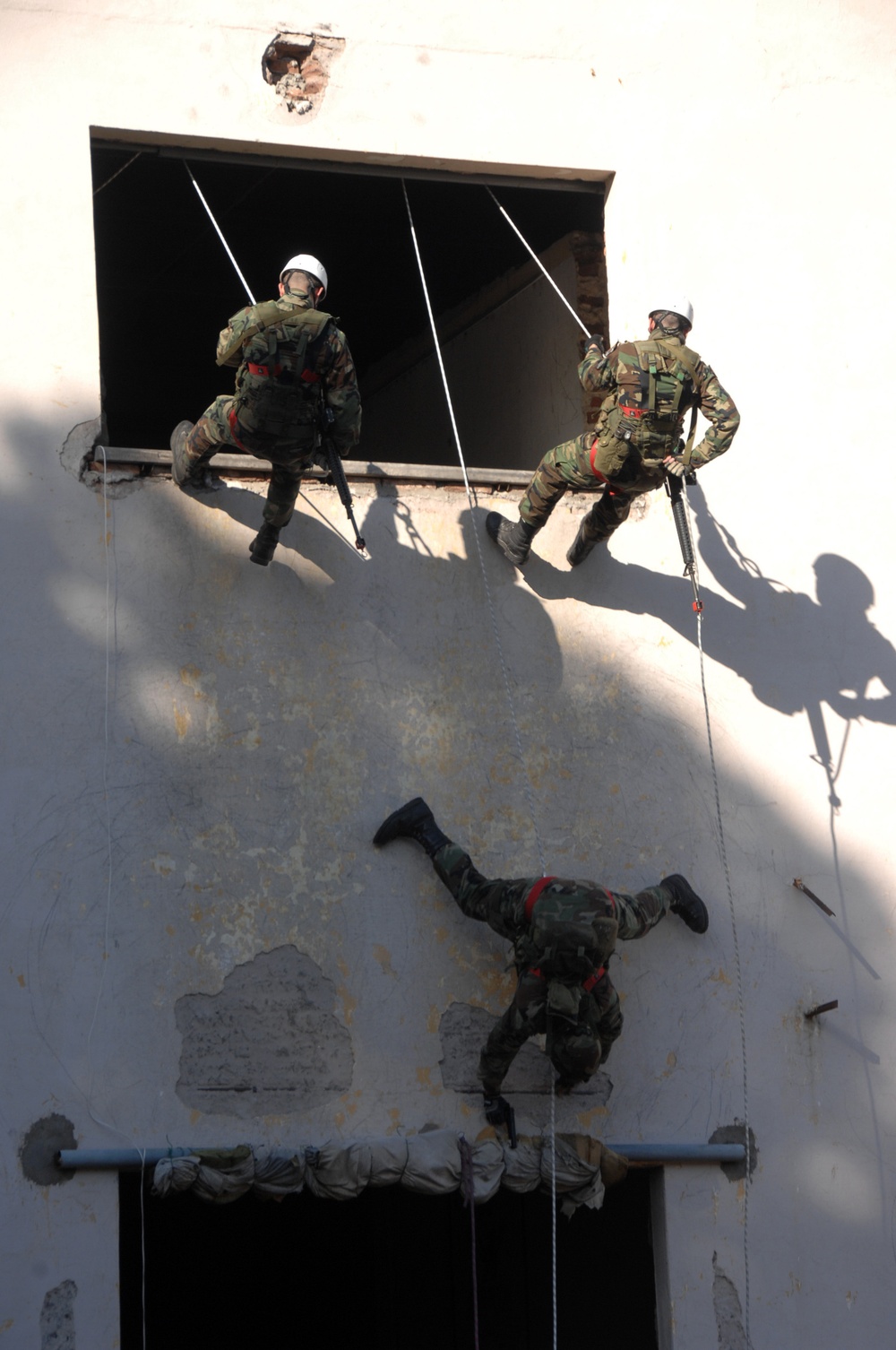 Three military personnel in camouflage uniforms are conducting a fast-rope or rappelling exercise on the exterior of a weathered white building. Two soldiers are positioned at an upper window opening while a third is mid-descent on the building's facade using ropes. The scene appears to be a tactical training exercise involving building assault or entry techniques.