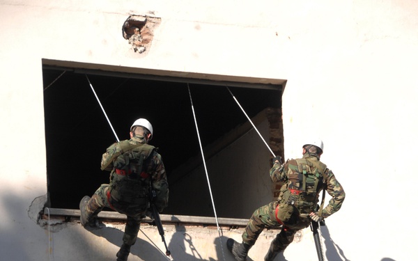 Three military personnel in camouflage uniforms are conducting a fast-rope or rappelling exercise on the exterior of a weathered white building. Two soldiers are positioned at an upper window opening while a third is mid-descent on the building's facade using ropes. The scene appears to be a tactical training exercise involving building assault or entry techniques.