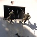 Three military personnel in camouflage uniforms are conducting a fast-rope or rappelling exercise on the exterior of a weathered white building. Two soldiers are positioned at an upper window opening while a third is mid-descent on the building's facade using ropes. The scene appears to be a tactical training exercise involving building assault or entry techniques.