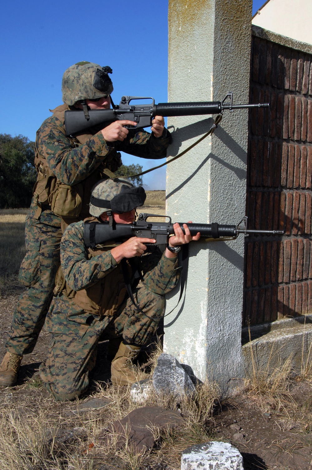 Two military personnel in MARPAT (Marine digital camouflage) uniforms are taking up firing positions at the corner of a stucco and brick building. One soldier is standing while the other is kneeling, both aiming M16-style rifles around the building corner. The setting appears to be a training exercise in an open, dry field environment.