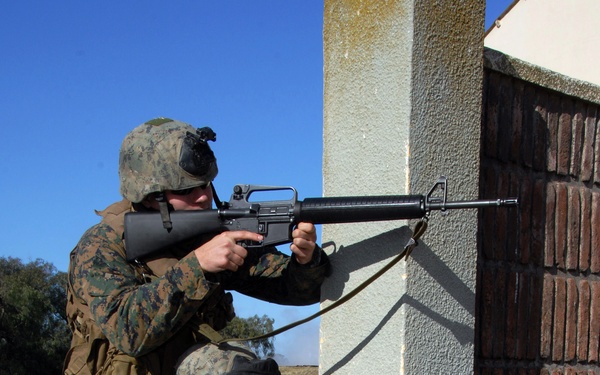 Two military personnel in MARPAT (Marine digital camouflage) uniforms are taking up firing positions at the corner of a stucco and brick building. One soldier is standing while the other is kneeling, both aiming M16-style rifles around the building corner. The setting appears to be a training exercise in an open, dry field environment.