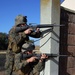 Two military personnel in MARPAT (Marine digital camouflage) uniforms are taking up firing positions at the corner of a stucco and brick building. One soldier is standing while the other is kneeling, both aiming M16-style rifles around the building corner. The setting appears to be a training exercise in an open, dry field environment.