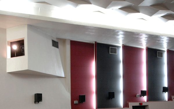 A military briefing or all-hands meeting is taking place in a large auditorium or theater. A senior naval officer in khaki uniform is speaking at a podium/stage with a microphone, addressing a large audience of military personnel in various uniforms including camouflage and blue uniforms. The venue appears to be a base theater or conference hall with decorative wall panels in red and gray.