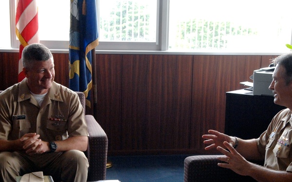 Two U.S. Navy officers in tan/khaki service uniforms are seated across from each other in what appears to be a military office, engaged in a formal meeting or discussion. Both wear ribbons and insignia on their uniforms, suggesting senior rank. The setting includes an American flag, a second flag (possibly a unit or state flag), and a dark wood-paneled office environment.