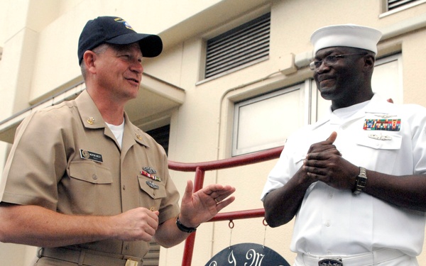 Two U.S. Navy personnel are engaged in conversation outside a building at what appears to be a naval installation in Atsugi, Japan. One is wearing khaki service uniform with a navy cap, and the other is in white dress uniform with multiple service ribbons. A sign reading 'MCPON' is visible in the background, along with an AIMD (Aircraft Intermediate Maintenance Department) unit insignia plaque.