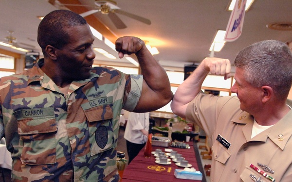 Two U.S. Navy personnel are engaged in a lighthearted interaction, both flexing their biceps and comparing muscles in what appears to be a dining facility or event hall. The individual on the left wears woodland camouflage BDUs with a name tape reading 'CANNON' and U.S. Navy insignia, while the individual on the right wears the tan/khaki Navy service uniform with multiple ribbons and badges. The setting appears to be a military dining or event facility with tables set up in the background.