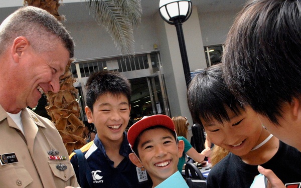A U.S. Navy officer in tan dress uniform is interacting cheerfully with a group of young boys, likely of Asian descent, in an outdoor public setting. The children are smiling and laughing while holding papers or notebooks, suggesting an autograph or meet-and-greet interaction. The atmosphere is warm and positive, indicating a community outreach or public relations event.