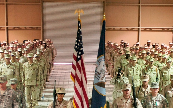 A large military formation ceremony is taking place inside what appears to be a gymnasium or large indoor facility. Hundreds of soldiers in desert camouflage uniforms are standing at attention in organized rows, with a color guard presenting the American flag and what appears to be a unit or command flag at the center. A podium is positioned in the foreground, suggesting a formal ceremony such as a change of command, deployment, or awards ceremony.