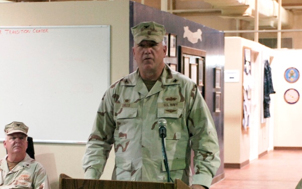 A U.S. Navy officer in desert camouflage (DCU) uniform stands at a wooden podium with a microphone, apparently delivering a speech or briefing. The setting is a 'Warrior Transition Center' as indicated by text on the whiteboard behind him. Two other military personnel in similar desert camouflage uniforms are seated on either side in the background.