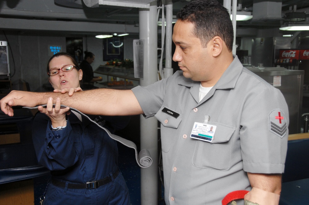 A medical training scenario aboard what appears to be a military vessel or facility, where a person in gray uniform with a Red Cross patch and name tag 'VIEIRA' is demonstrating or practicing a tourniquet application technique on their own arm. A second person in dark blue uniform wearing glasses is observing and assisting with the procedure. The setting appears to be a common area or mess hall of a military ship or installation.