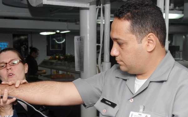 A medical training scenario aboard what appears to be a military vessel or facility, where a person in gray uniform with a Red Cross patch and name tag 'VIEIRA' is demonstrating or practicing a tourniquet application technique on their own arm. A second person in dark blue uniform wearing glasses is observing and assisting with the procedure. The setting appears to be a common area or mess hall of a military ship or installation.