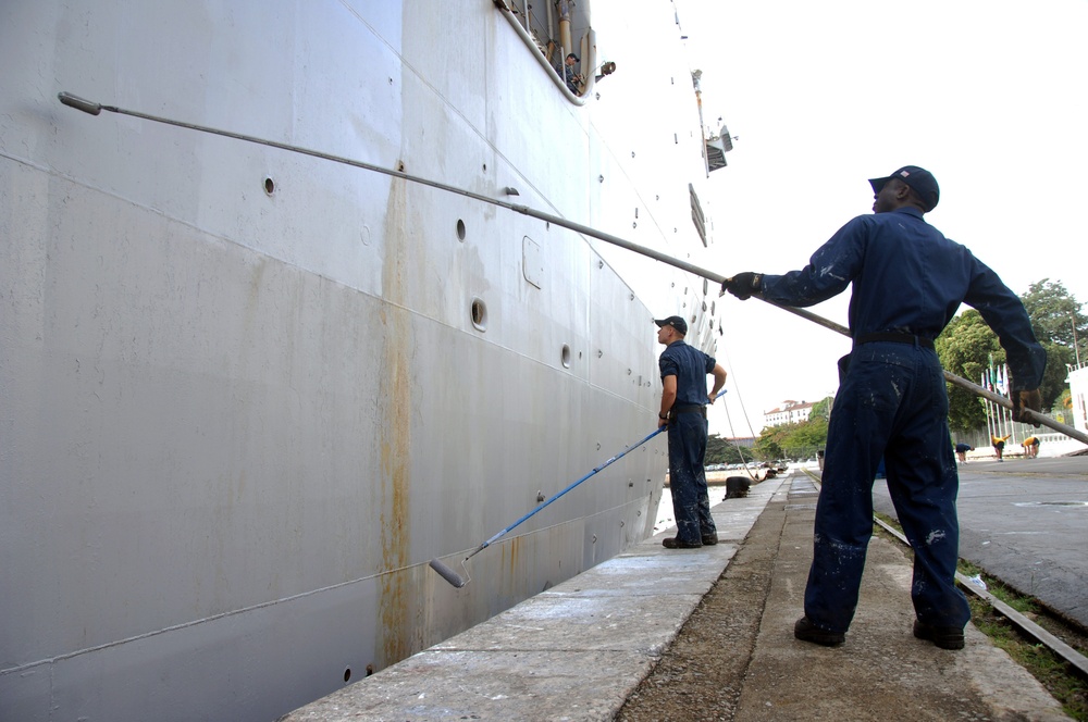Two naval personnel in dark blue uniforms are performing maintenance work on the hull of a large gray military vessel docked at a pier. One worker is using a long-handled paint roller to apply paint to the ship's hull, while the other holds an extended pole tool. The ship is moored at what appears to be a port facility with trees and buildings visible in the background.