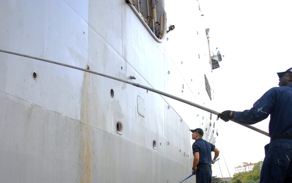 Two naval personnel in dark blue uniforms are performing maintenance work on the hull of a large gray military vessel docked at a pier. One worker is using a long-handled paint roller to apply paint to the ship's hull, while the other holds an extended pole tool. The ship is moored at what appears to be a port facility with trees and buildings visible in the background.