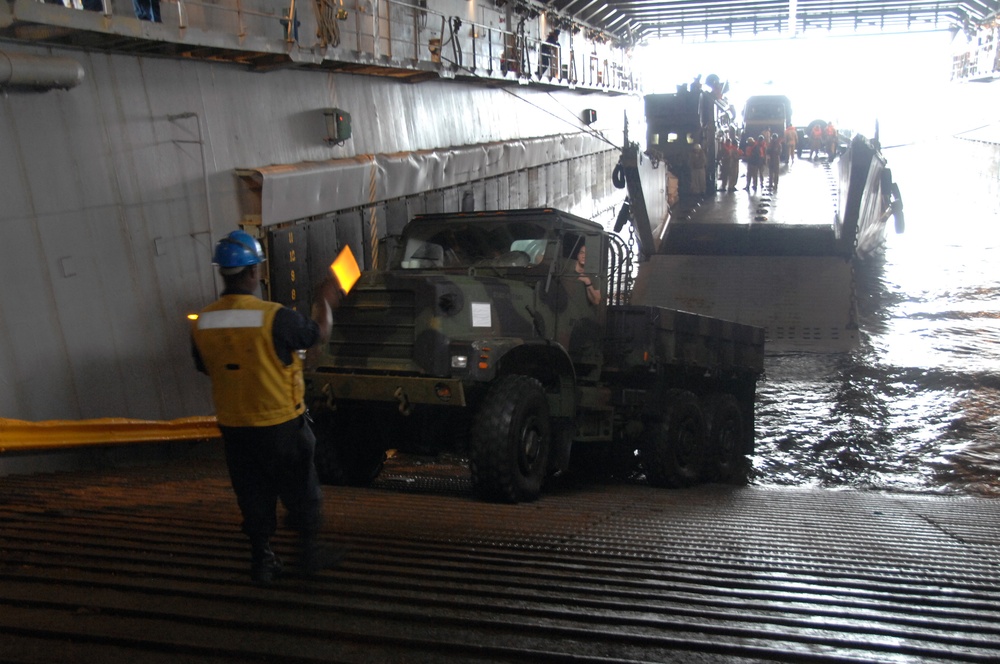 A military cargo loading operation is underway inside what appears to be the well deck of a naval amphibious ship. A camouflage-painted military cargo truck is being guided by a deck handler wearing a yellow safety vest and blue hard hat, who is using a signal wand. Additional military vehicles and personnel in orange vests are visible in the background near the open stern ramp.