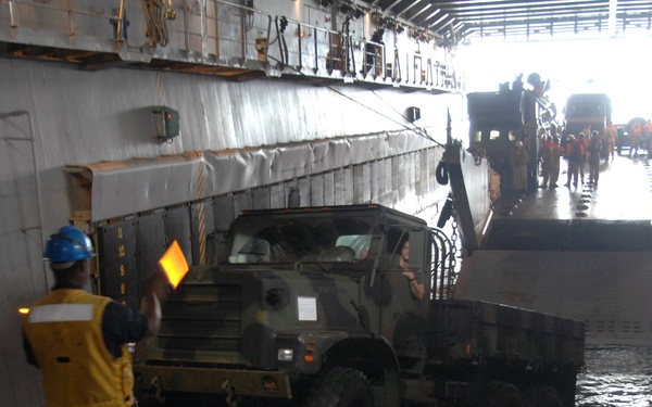 A military cargo loading operation is underway inside what appears to be the well deck of a naval amphibious ship. A camouflage-painted military cargo truck is being guided by a deck handler wearing a yellow safety vest and blue hard hat, who is using a signal wand. Additional military vehicles and personnel in orange vests are visible in the background near the open stern ramp.