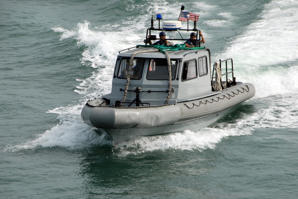 A gray military or law enforcement rigid inflatable boat (RIB) is underway at speed on open water, creating a significant wake. The vessel is equipped with navigation equipment, a blue emergency light, and an American flag mounted on top. Two personnel are visible on the upper deck/gun platform area of the boat.
