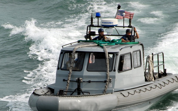 A gray military or law enforcement rigid inflatable boat (RIB) is underway at speed on open water, creating a significant wake. The vessel is equipped with navigation equipment, a blue emergency light, and an American flag mounted on top. Two personnel are visible on the upper deck/gun platform area of the boat.