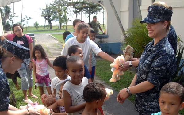 Two U.S. Navy personnel in digital camouflage uniforms are distributing stuffed teddy bears to a group of children in what appears to be a humanitarian outreach or community relations event. The setting appears to be an outdoor area near a building in a tropical location, with palm trees visible in the background. Several children are already holding teddy bears while others wait in line to receive them.