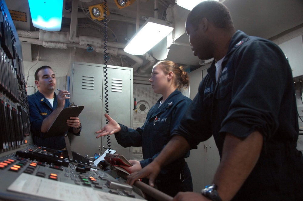 Three U.S. Navy personnel in dark blue uniforms are gathered around a control panel in what appears to be a shipboard operations room or engineering space. A female sailor in the center appears to be briefing or explaining something to her colleagues, one of whom holds a clipboard. The room contains various control panels with illuminated buttons, a wall-mounted telephone, and overhead lighting.