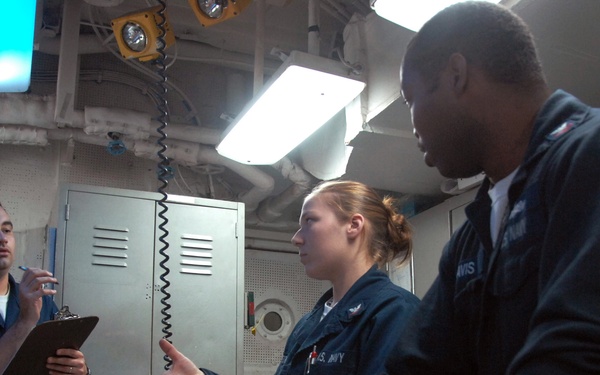 Three U.S. Navy personnel in dark blue uniforms are gathered around a control panel in what appears to be a shipboard operations room or engineering space. A female sailor in the center appears to be briefing or explaining something to her colleagues, one of whom holds a clipboard. The room contains various control panels with illuminated buttons, a wall-mounted telephone, and overhead lighting.