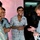 A Western man in a light blue button-down shirt is engaged in conversation with three younger men near a pink-painted wall, likely outside a small shop or building. The interaction appears friendly and animated, with at least two of the men smiling or laughing. The Western man is holding papers and gesturing with his hand, suggesting an interview, survey, or informal discussion.