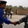 An Iraqi Police officer in a blue uniform shakes hands with a U.S. Army soldier in ACU (Army Combat Uniform) camouflage. The Iraqi officer is holding two water bottles while greeting the soldier, suggesting a cooperative interaction. The setting appears to be a checkpoint or forward operating base in Iraq, with palm trees, sandbag fortifications, and concertina wire visible in the background.