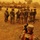 A group of approximately 12-15 soldiers in US Army ACU (Army Combat Uniform) digital camouflage are gathered in a briefing circle on a gravel surface during a severe sandstorm, giving the scene a heavy orange-yellow cast. A soldier in the foreground, with the name tape 'LONGBINE' visible, is filming the group with a video camera. Multiple military vehicles including MRAPs and Humvees are staged in the background.