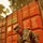 A soldier in US Army ACU (Army Combat Uniform) camouflage is crouching on a gravel surface while handling a professional video camera near ground level. The soldier is positioned in front of large red intermodal shipping containers marked with weight specifications and container identification codes. The setting appears to be a military logistics or forward operating base area.