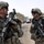 Two U.S. Army soldiers in full combat gear are engaged in a casual conversation, both smiling, in what appears to be a Middle Eastern urban or semi-urban environment. Both soldiers are equipped with assault rifles, body armor, helmets, and tactical accessories. The background shows palm trees, vehicles, and a roadside market area consistent with an operational deployment setting.