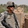 A U.S. Army soldier identified by name tape as 'LAMMERT' stands at what appears to be a fortified checkpoint or patrol position. The soldier is in full combat gear including ACU (Army Combat Uniform) digital camouflage, body armor, helmet with NVG mount, and is carrying an M249 Squad Automatic Weapon (SAW). In the background, a tan-colored MRAP (Mine-Resistant Ambush Protected) vehicle is visible alongside palm trees and HESCO barrier fortifications, consistent with a Middle Eastern operational environment.
