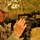 A female soldier in US Army ACU (Army Combat Uniform) digital camouflage is operating a professional Sony DV video camera mounted on a tripod. The setting appears to be an arid, dusty outdoor environment consistent with a Middle Eastern deployment location. The soldier appears to be performing military public affairs or combat camera documentation duties.