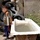 A young boy stands outdoors next to a worn, water-filled bathtub that has been repurposed outside. The setting appears to be a low-income or informal residential area, with discarded tires stacked against a concrete block wall in the background. The environment shows signs of poverty and improvised resource use.
