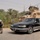 The image shows a military checkpoint or patrol operation on a road in what appears to be a Middle Eastern environment, likely Iraq, based on the vegetation, terrain, and Arabic license plate visible on the civilian vehicle. A U.S. soldier in full combat gear is taking a crouched tactical position beside a black civilian sedan, while a tan MRAP (Mine-Resistant Ambush Protected) military vehicle is positioned in the background. The scene suggests a vehicle stop or security check operation.