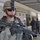 A U.S. Army soldier in full combat gear stands in the foreground holding an M4 carbine rifle at a ready position. The setting appears to be a commercial street or market area with shuttered storefronts and Arabic-language signage. In the background, several civilians and what appears to be a local security officer are conversing near a building entrance.
