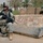 A U.S. Army soldier in full combat gear crouches beside a concrete barrier with Arabic script painted in red on it. The setting appears to be a checkpoint or fortified position, evidenced by sandbags and HESCO-style barriers in the background. Palm trees and dense vegetation are visible in the background, consistent with a Middle Eastern environment.