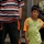 A young boy wearing a green and orange 'sdiboy' branded t-shirt stands next to an adult whose torso is visible, wearing a striped shirt and dark athletic pants. The boy appears to be looking directly at the camera with a neutral expression. The setting appears to be an outdoor or semi-covered public area with blurred figures visible in the background.