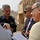 The image shows a group of four men engaged in a discussion outdoors near a military armored vehicle. One man is wearing an Iraqi Police uniform with Arabic text reading 'Ministry of Interior' and an 'IP' (Iraqi Police) patch. A notepad with handwritten notes is visible, suggesting a formal meeting or briefing is taking place.